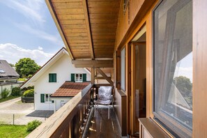 Un balcón de madera con barandilla de metal, vista a una casa vecina con techo de tejas y cielo despejado.
