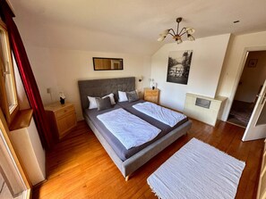 Quarto com cama de casal, mesa de cabeceira, abajur, espelho, radiador e porta que dá para outro cômodo.