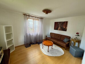 A living room with a sofa, a coffee table, a bookshelf, a window with sheer curtains, and a painting on the wall.