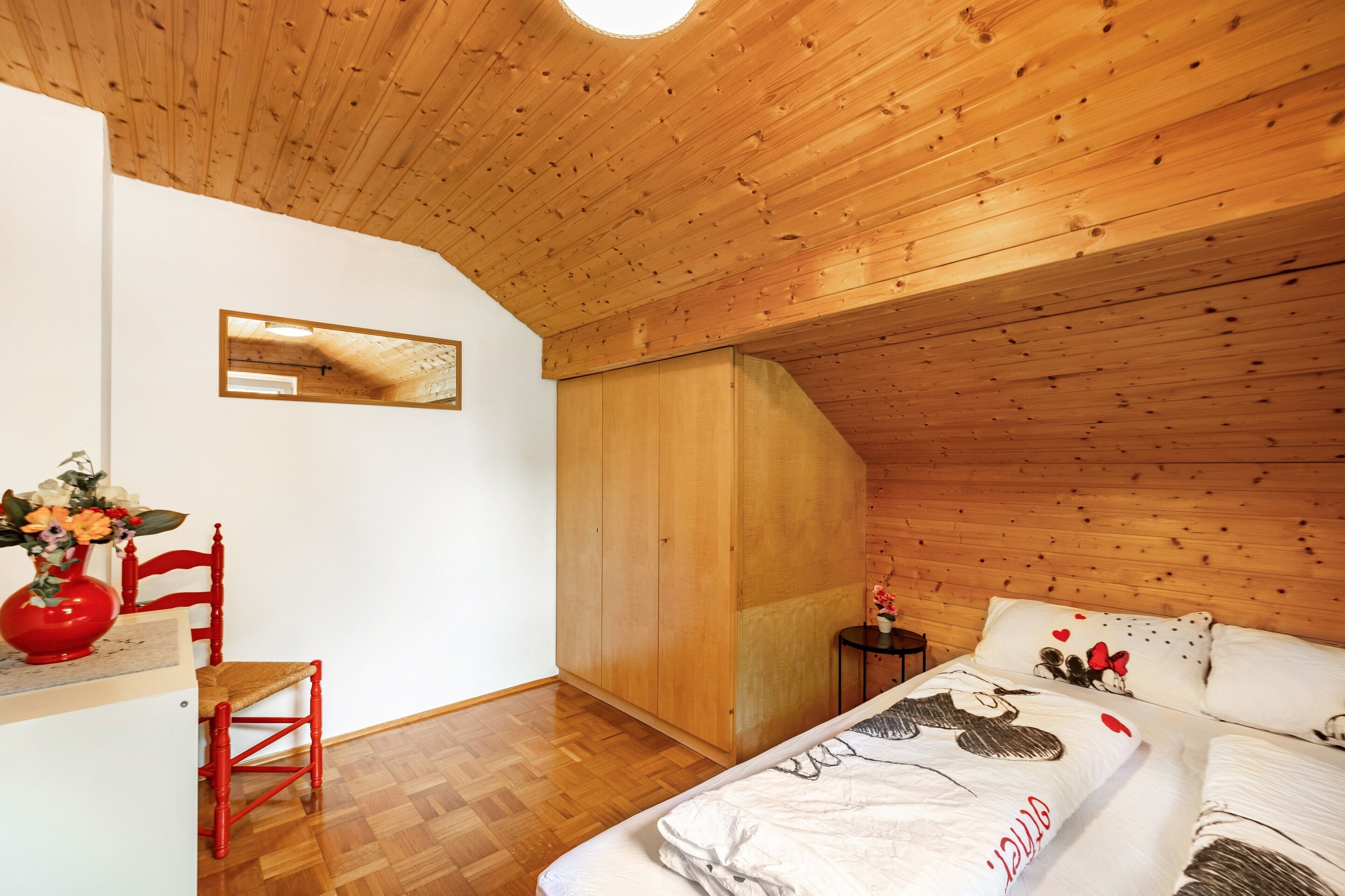 Ein Schlafzimmer mit Holzbalkendecke, einem Bett mit weiß-rotem Dekor, einem roten Sessel, einem kleinen Tisch mit einer Vase und einem Kleiderschrank.