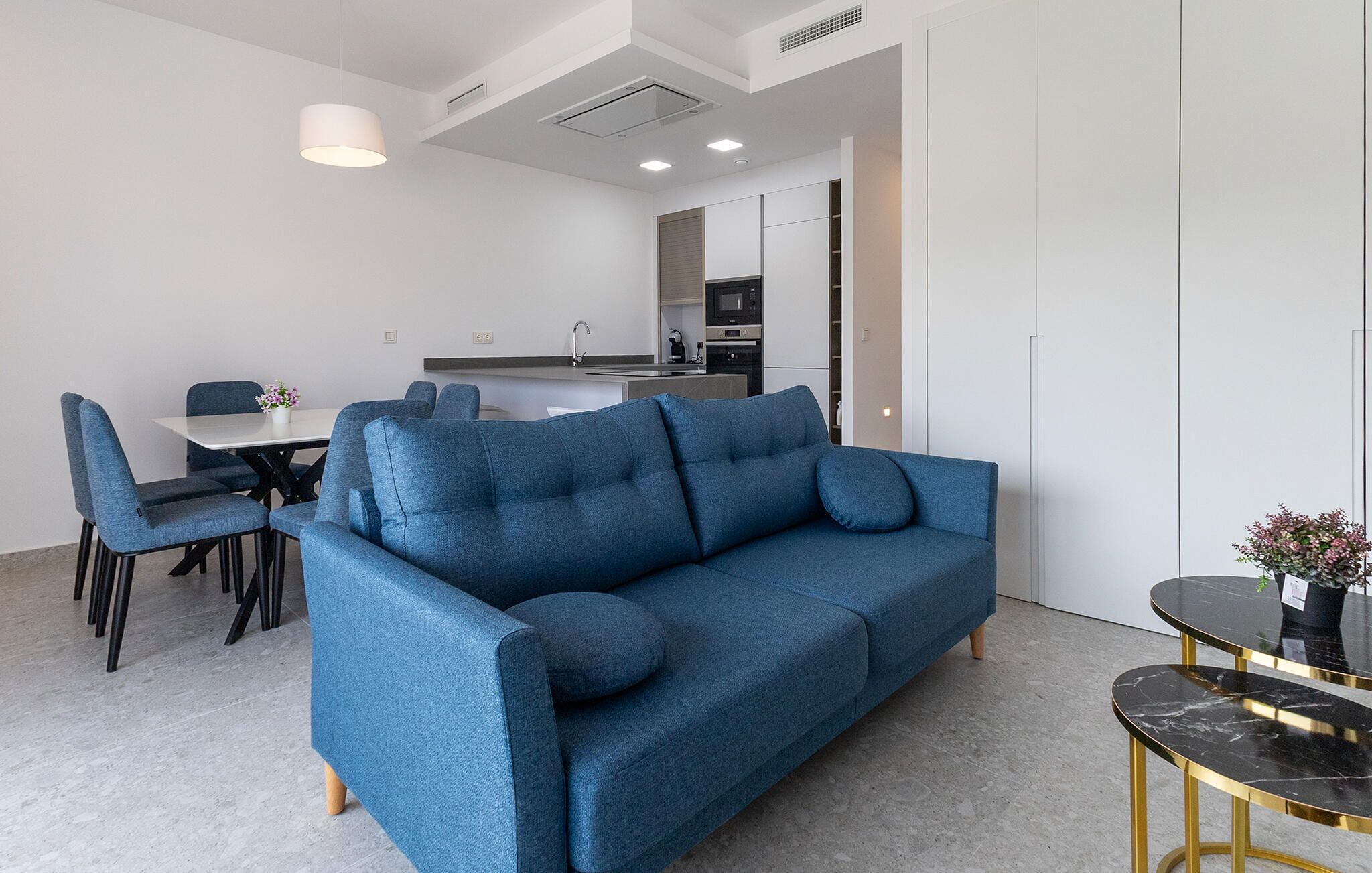 En moderne stue med en blå sofa, et spisebord med stole, et køkkenområde og et lille sofabord med en vase.