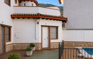 Ein zweistöckiges Gebäude mit weißer Fassade, einem Ziegeldach und einem steinernen Sockel. Es gibt eine Tür mit Fenster, einen kleinen Pool und Kübelpflanzen.