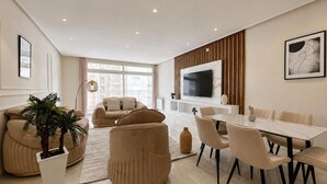 Ein modernes Wohnzimmer mit einer Sofagarnitur, einem Esstisch mit Stühlen, einem an der Wand montierten Fernseher und einem großen Fenster, das für natürliches Licht sorgt.