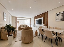 Una sala moderna con un conjunto de sofá, una mesa de comedor con sillas, un televisor montado en la pared y un gran ventanal que proporciona luz natural.