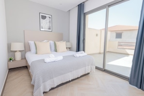 A modern hotel room with a large bed, a nightstand with a lamp, a small plant, and a view through a sliding glass door.