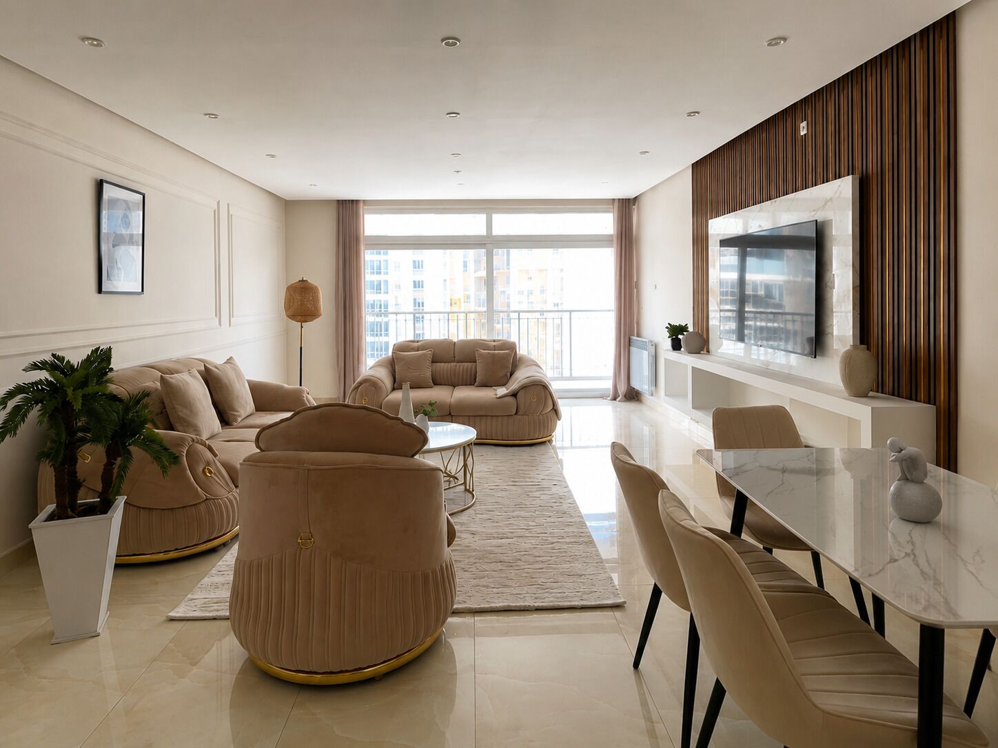 A modern living room with a sofa set, a dining table with chairs, a TV mounted on the wall, and a large window providing natural light.