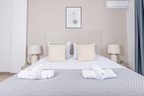 A neatly made bed with two pillows, two bedside lamps, a framed artwork, and a wall-mounted air conditioner.