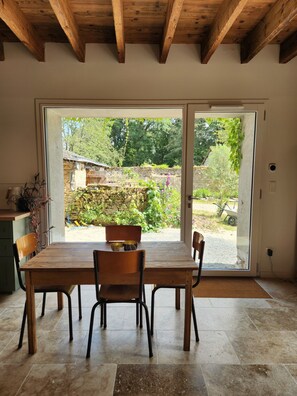 A dining area with a wooden table and chairs, a view of a garden through a glass door, and a stone wall in the background. - Tastefully renovated 18th century farmhouse, landscaped gardens and access to the château grounds (Treillières)