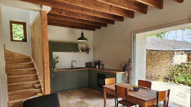 A kitchen with wooden beams, a dining area with a table and chairs, and a view of a stone building outside. - Tastefully renovated 18th century farmhouse, landscaped gardens and access to the château grounds (Treillières)
