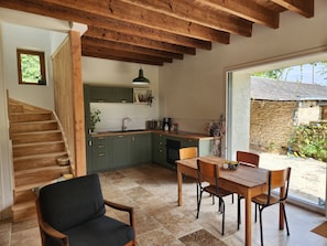 A kitchen with wooden beams, a dining area with a table and chairs, and a view of a stone building outside. - Tastefully renovated 18th century farmhouse, landscaped gardens and access to the château grounds (Treillières)