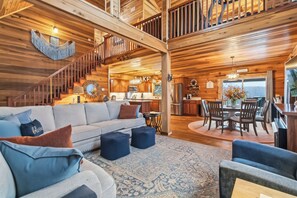 Un vaste espace de vie comprenant un canapé, des poufs et une table à manger avec des chaises. On peut apercevoir une cuisine avec des meubles en bois et un escalier menant à un étage supérieur.