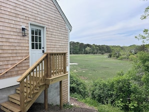 A wooden house with a white door, a wooden staircase, and a view of a green field. - Newly Renovated Marshview Retreat in the Heart of Wellfleet (2440) (Wellfleet)