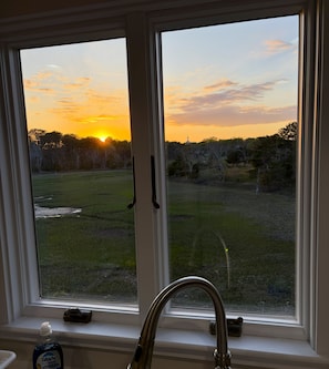 A window with a view of a sunset over a grassy field, a metal faucet, and a bottle of Dawn Platinum dish soap on the windowsill. - Newly Renovated Marshview Retreat in the Heart of Wellfleet (2440) (Wellfleet)