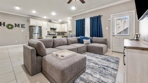 Un salon moderne avec un canapé d’angle, un ventilateur de plafond et une cuisine en arrière-plan.