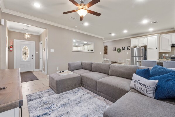 A modern living room with a sectional sofa, a dining table, and a kitchen area in the background. - 3 Bedroom Duplex - The Gardens Cottage Duo A (Houston)