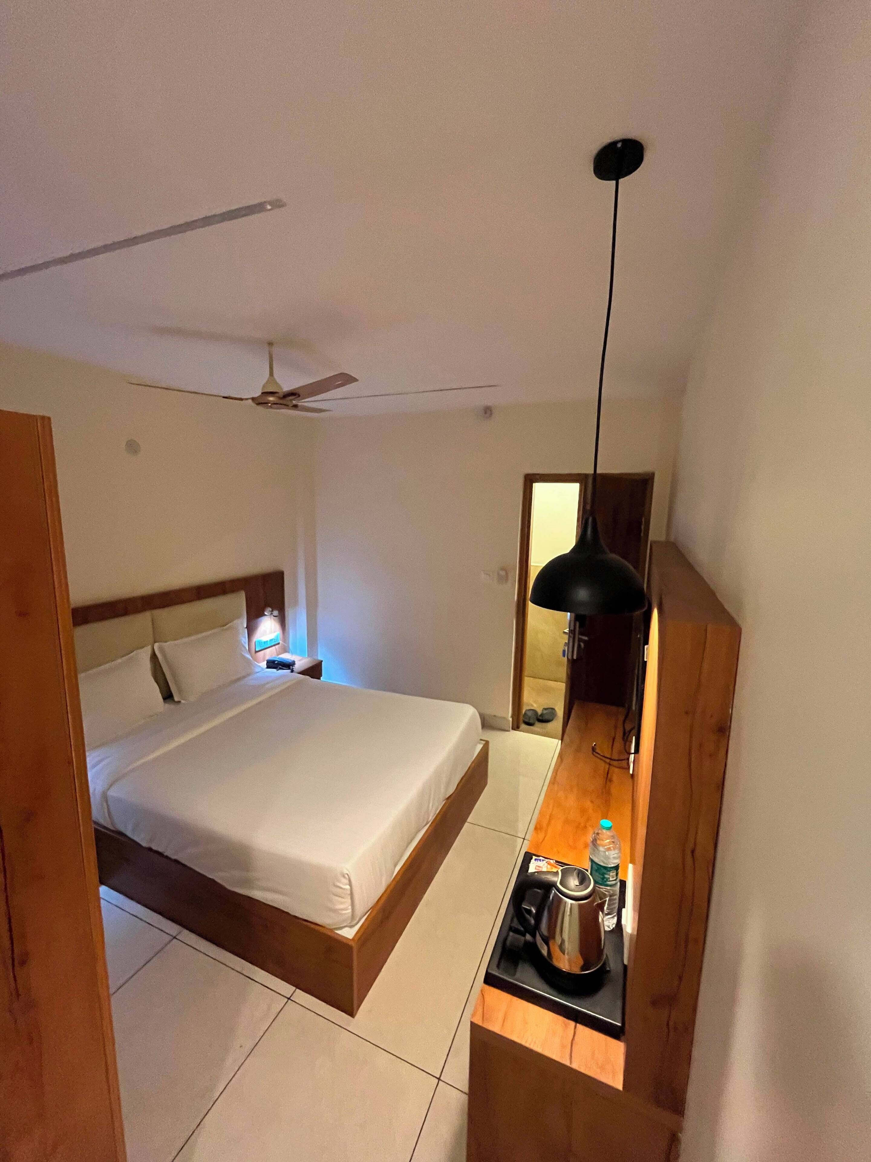 Un dormitorio con una cama, una mesita de noche, un ventilador de techo, un escritorio de madera con una tetera y un espejo.