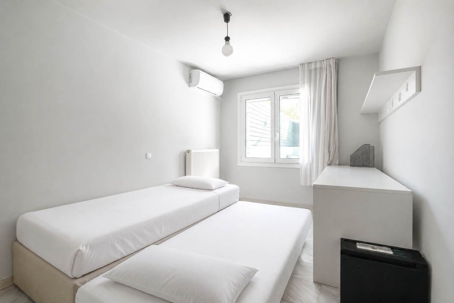 Una habitación minimalista con dos camas individuales, un radiador, una ventana con cortinas, un escritorio y un cubo de basura.