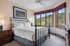 Ein Schlafzimmer mit Bett, Nachttisch, Lampe, Deckenventilator und großen Fenstern mit Blick auf Grünflächen.