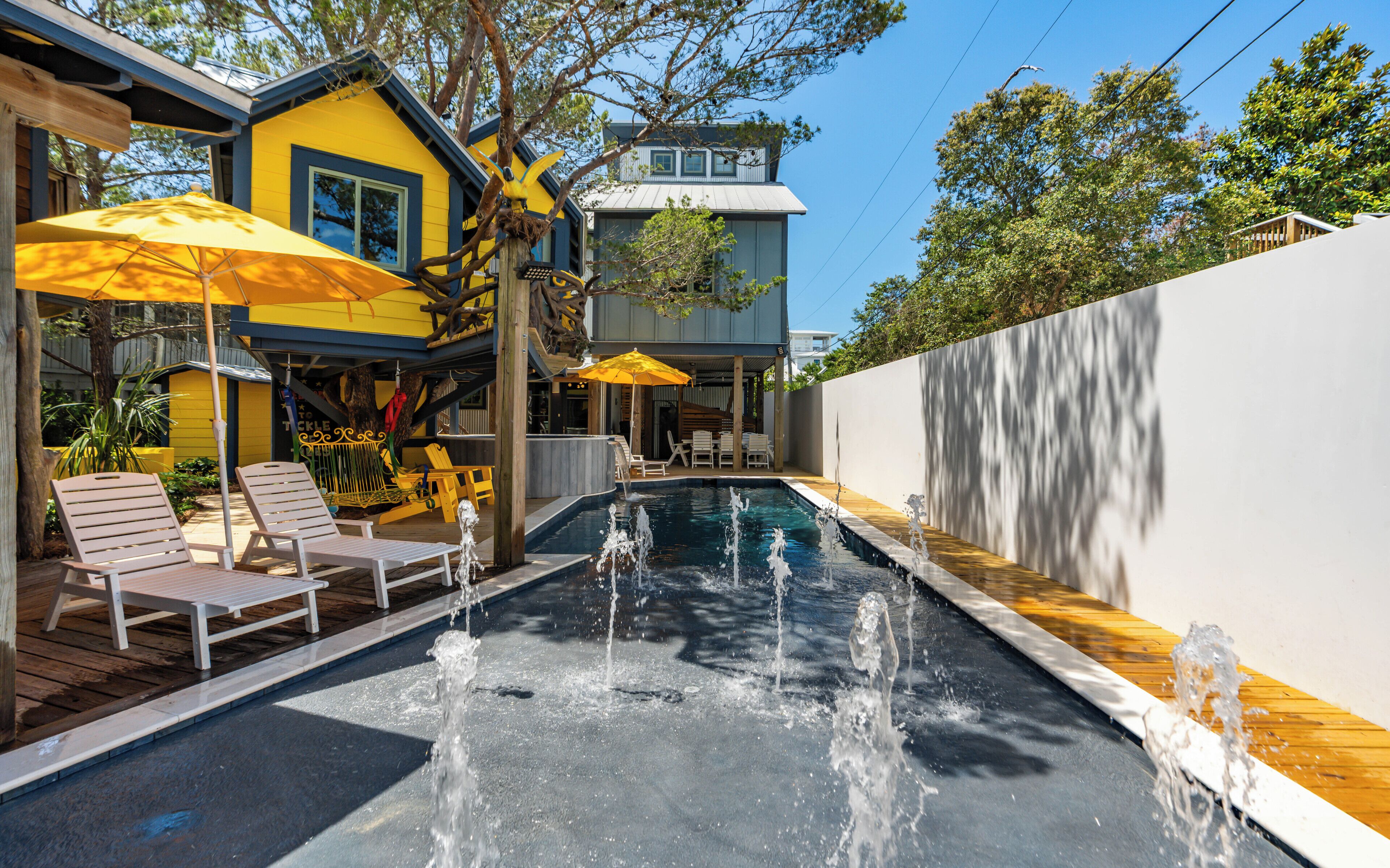 A swimming pool with water fountains, outdoor seating, and a yellow building with a blue roof.