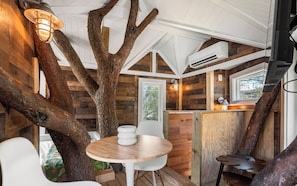 Ein rustikales Cabin-Interieur mit einem großen Baumstamm, einem runden Holztisch, weißen Stühlen und einem Fenster mit Blick auf viel Grün.