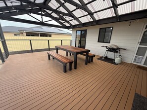 A covered outdoor area with a wooden deck, two wooden tables, a bench, and a barbecue grill.