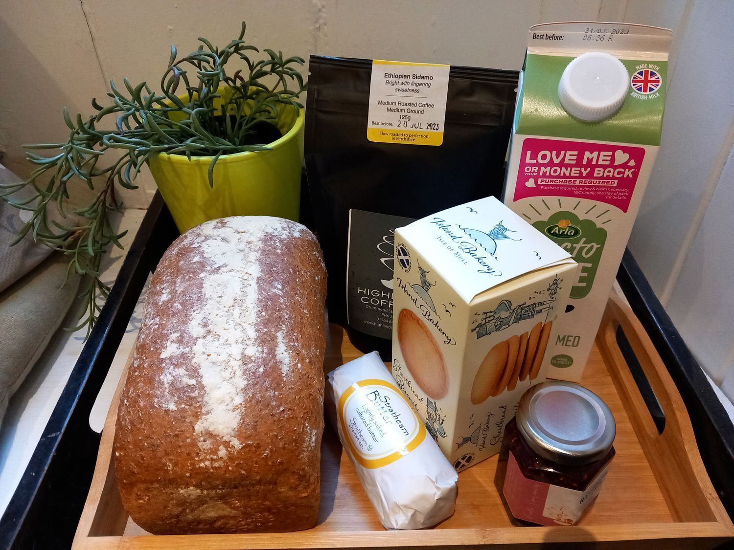 Un plateau contenant un pain, une plante, un paquet de café, un carton de lait, un pot de confiture et un paquet de beurre.