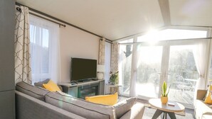 A modern living room with a sofa, a television, a small table with a plant, and a balcony with outdoor seating.