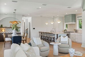 A modern kitchen with white cabinetry, a dining area with a round table and chairs, and a living room with sofas and a coffee table. - Hardin Hideaway - Anna Maria Island (Anna Maria)
