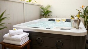 A hot tub with two glasses of white wine, a bucket with grapes and a bottle, and a stack of white towels on a wooden stool.