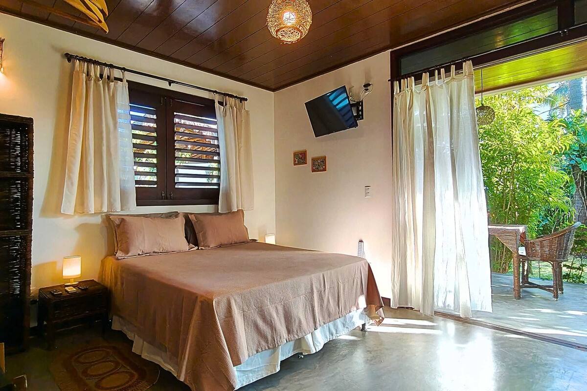 A bedroom with a bed, curtains, a television, and a view of greenery outside.