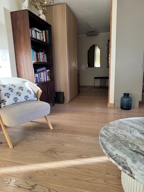 Un salon doté d’un plancher en bois, d’une table ronde en marbre, d’une bibliothèque et d’un fauteuil agrémenté d’un coussin à motifs floraux.