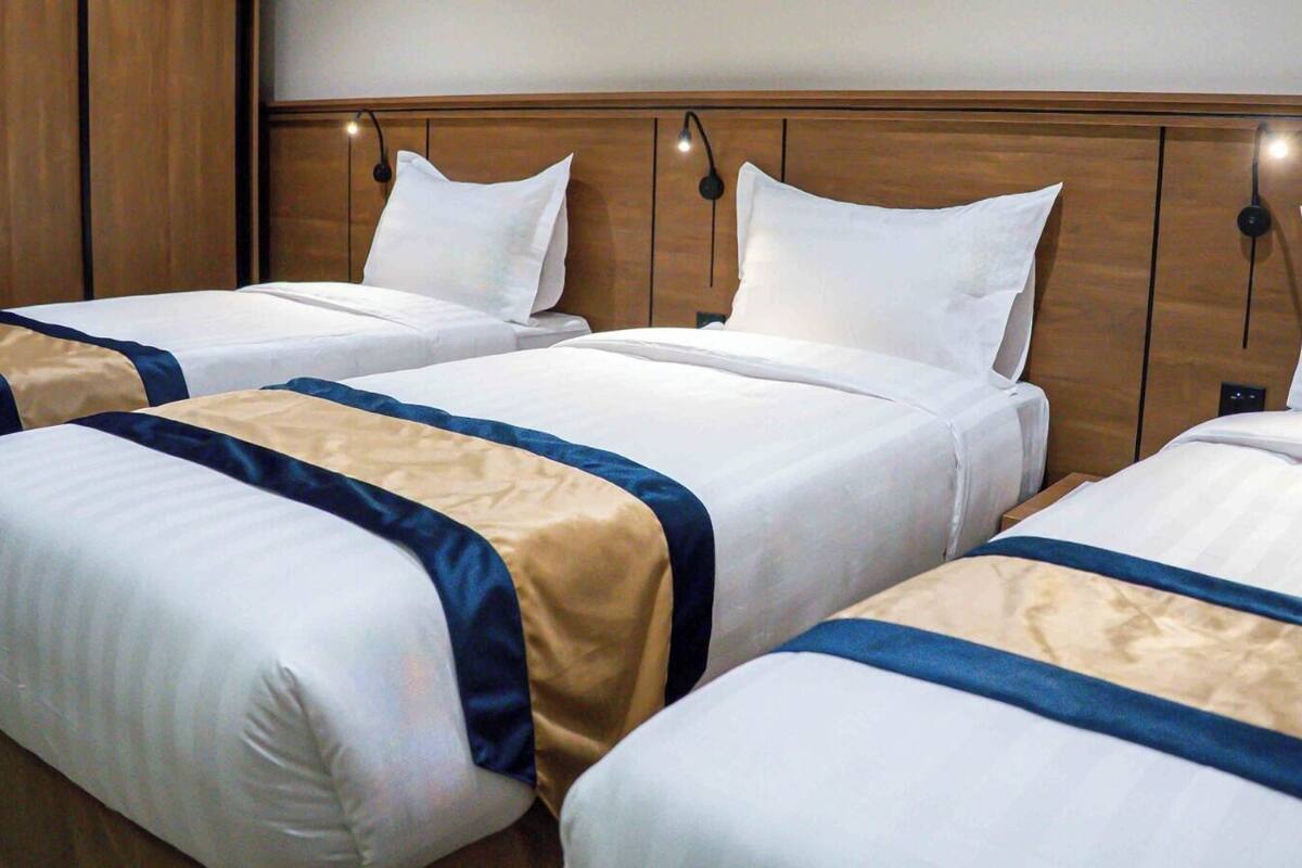 Tres camas individuales con sábanas blancas y colchas azules y beige, cabeceras de madera y lámparas de lectura fijadas a la pared.