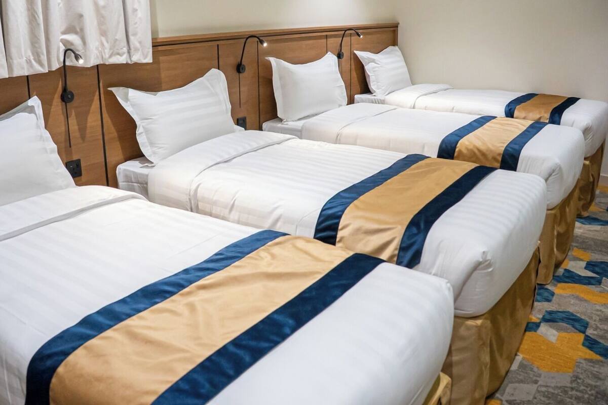 Habitación de hotel con cuatro camas dispuestas en fila, cada una con ropa blanca y detalles en azul y beige.