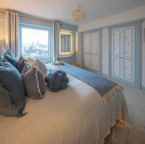 Ein Schlafzimmer mit einem großen Bett, Blick auf die Stadt durch ein Fenster, einer Kristallleuchter und einer Wand mit gerahmten Bildern.
