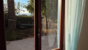 Een kamer met een groot raam, een houten deur en uitzicht op een buitenruimte met een hangmat, een terras en bomen.