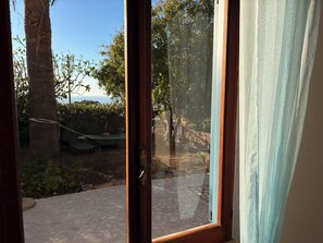 A room with a large window, a wooden door, and a view of an outdoor area with a hammock, a stone patio, and trees. - Apartment in a villa under a large palm tree and garden (Porto Alabe)