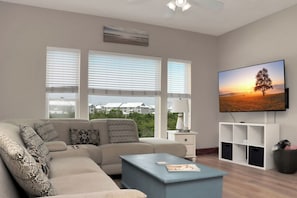A living room with a sofa, coffee table, TV, and a view of houses through large windows. - 🌴 Short Walk to Beach - Pet Friendly - Living the Dream ☀️ (Mexico Beach)