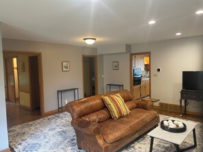 A living room with a brown leather sofa, a coffee table with decorative items, a television, and a kitchen visible through an open doorway. - Peaceful 2-bedroom brick Shorewood house near Lake Michigan beaches. (Shorewood)