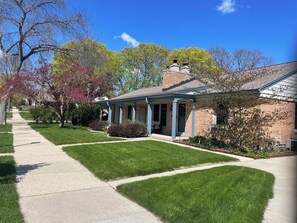 A residential house with a covered porch, a brick chimney, and a well-maintained lawn. - Peaceful 2-bedroom brick Shorewood house near Lake Michigan beaches. (Shorewood)