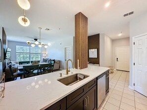 A modern kitchen with a central island, a built-in sink, and a dining area with a green table and chairs. - Beautiful Three Bed Condo, Minutes From Disney 3 Bedroom Condo (Kissimmee)