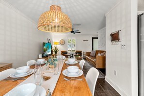 Ein Essbereich mit Holztisch, gedeckten Plätzen für vier Personen, weißen Stühlen, einer gewobenen Pendelleuchte und Blick in das Wohnzimmer mit Sofa und Sesseln.