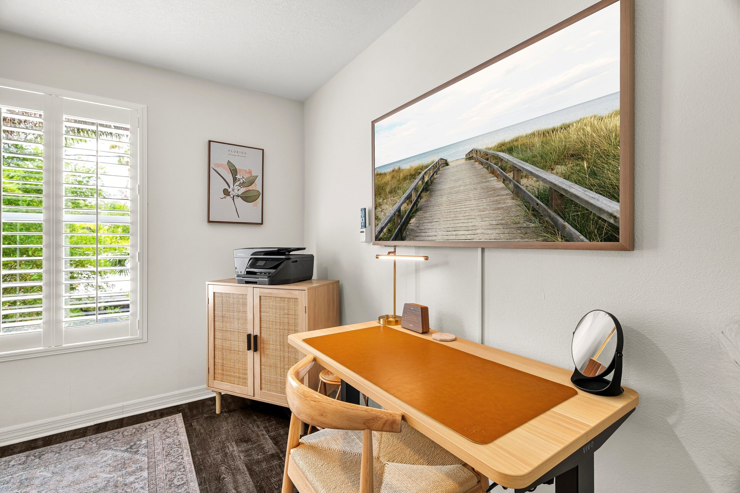 Une pièce comprenant un bureau en bois, une chaise, une lampe, une imprimante, un meuble, un tableau encadré et une grande fenêtre avec des stores.