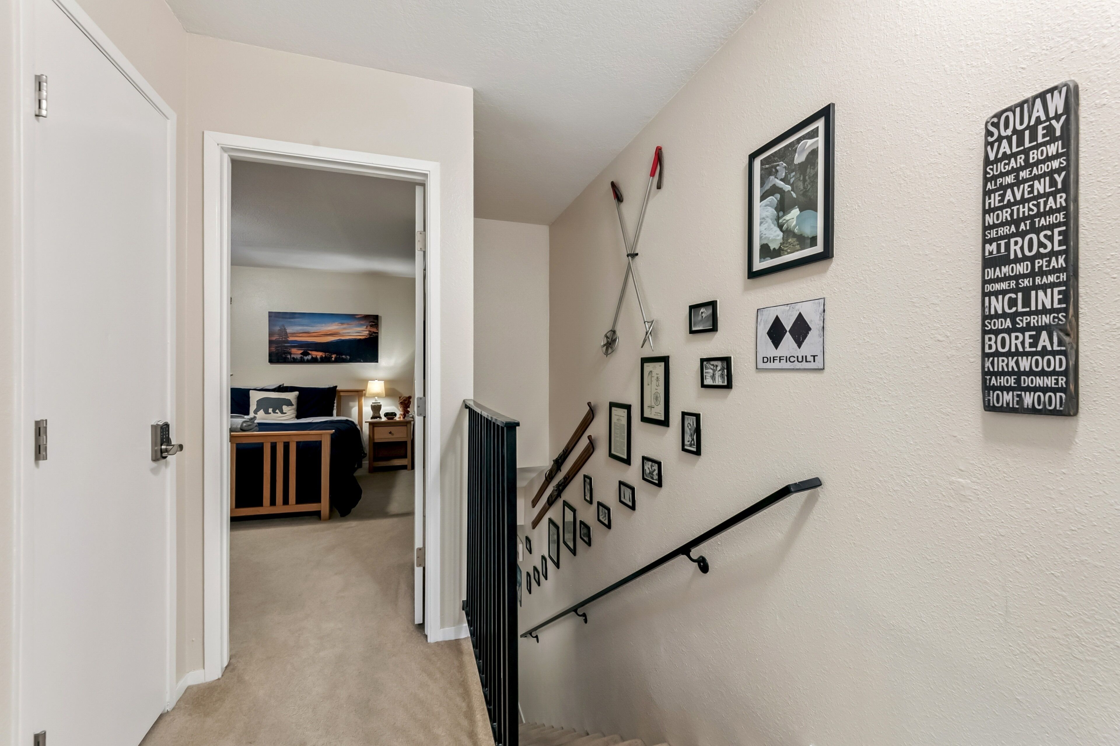 A hallway with a staircase, a door leading to a bedroom, and a wall with framed pictures and a sign listing ski locations.