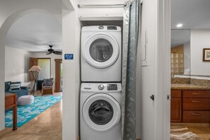 A laundry area with stacked washers and dryers, a blue patterned rug, a ceiling fan, and a view into a bathroom with a shower and sink. - Kauai Kahala At Poipu Kai 212 1 Bedroom Condo (Koloa)
