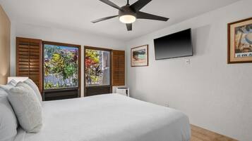 A bedroom with a bed, a ceiling fan, a TV, and a window with shutters.