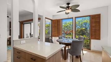 A modern kitchen with a central island, dining area with chairs, large windows with shutters, and a ceiling fan.