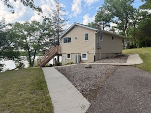 A two-story house with a wooden staircase leading to a deck, a gravel driveway, and a lake view.