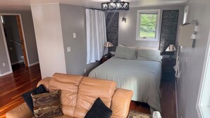 Quarto com cama, sofá de couro bege, mesa de cabeceira, abajur e janela.