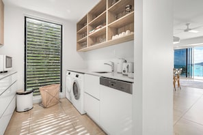 A modern kitchen with a built-in washer and dryer, a wooden shelf with kitchenware, and a large window with blinds. - Airlie Beach Penthouse (Airlie Beach)