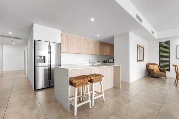 Une cuisine moderne avec un réfrigérateur en acier inoxydable, un meuble en bois et un plan de travail blanc. On trouve deux tabourets en osier près du bar.